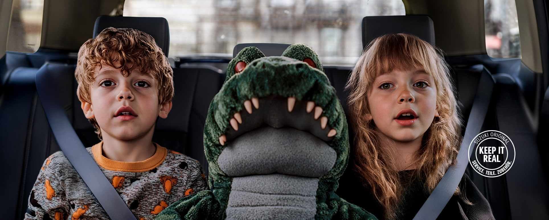 Zwei kleine Kinder sitzen angeschnallt auf einer Autorückbank, zwischen sich ein großes Plüschkrokodil, im unteren Bereich ist das Logo „Keep it real“ in weiß zu sehen