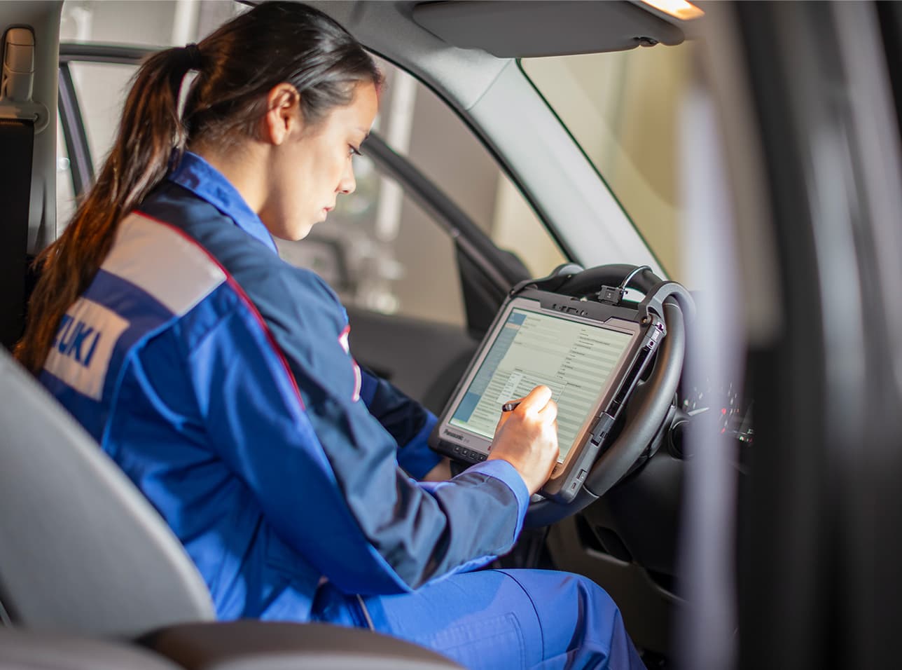 Eine Frau in einem blauen Suzuki-Arbeitsoverall sitzt mit Tablet hinter dem Lenkrad und wertet Daten aus