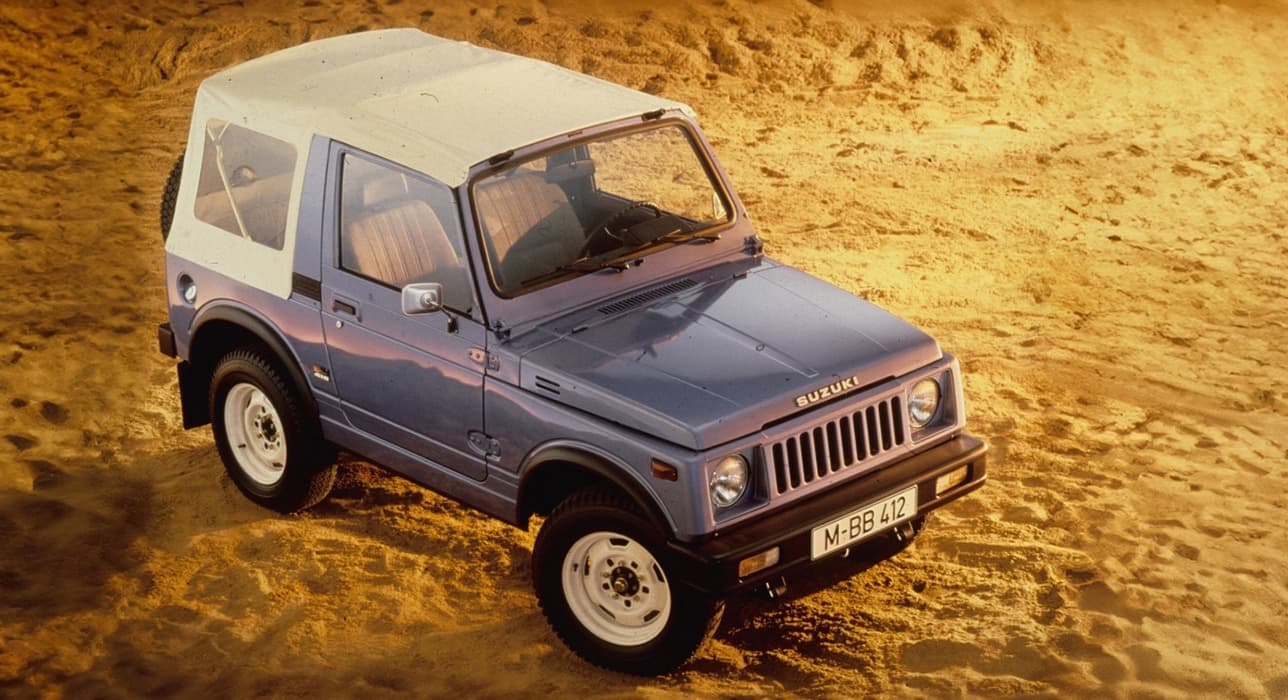 Abbildung zeigt von schräg oben einen blauen Jimny mit einem weißen Dach, der im Sand parkt