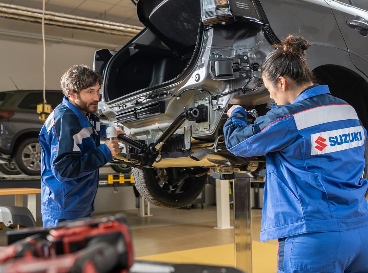 Ein Mann und eine Frau im blauen Suzuki-Arbeitsoutfit arbeiten an einem Auto, das mit der Hebebühne hochgefahren wurde