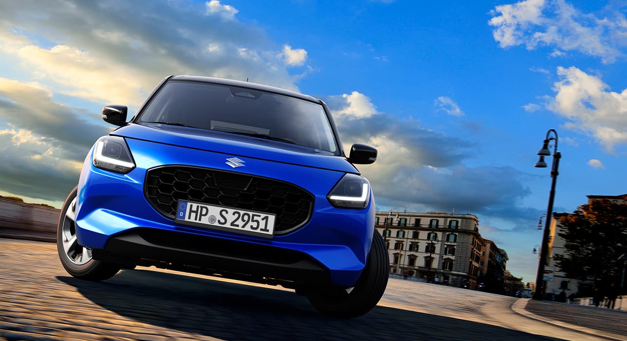 Ein blauer Suzuki Swift fährt in Frontalansicht um eine Kurve in städtischer Umgebung