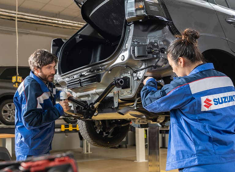 Ein Mann und eine Frau im blauen Suzuki-Arbeitsoutfit arbeiten an einem Auto, das mit der Hebebühne hochgefahren wurde