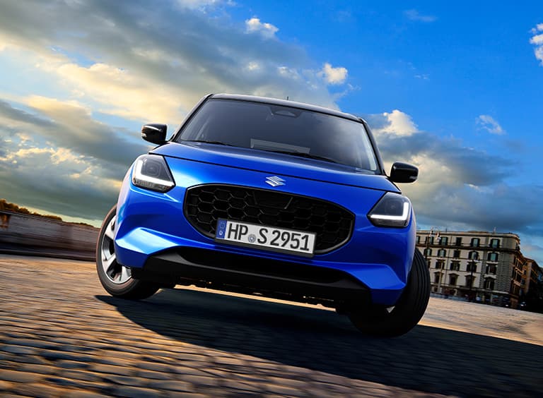 Ein blauer Suzuki Swift fährt in Frontalansicht um eine Kurve in städtischer Umgebung