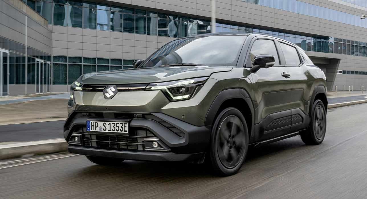 Ein dunkelgrüner e VITARA fährt auf einer Straße, im Hintergrund ist ein großes Gebäude mit Glasfront zu sehen.