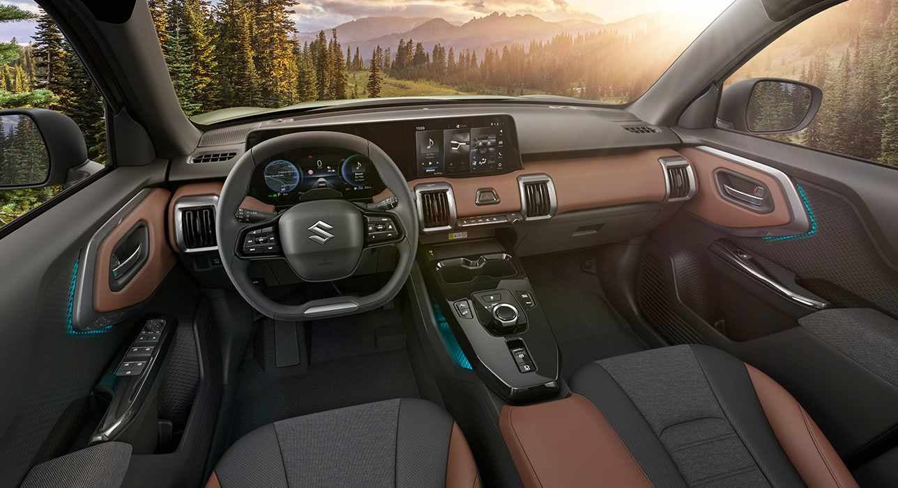 Der Innenraum des e VITARA vom Fahrersitz aus gesehen mit Fokus auf Lenkrad und Kontrollzentrum, durch die Frontscheibe sind ein sonnenbeschienener Wald und Berge zu sehen.