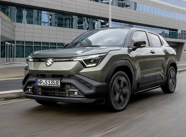 Ein dunkelgrüner e VITARA fährt auf einer Straße, im Hintergrund ist ein großes Gebäude mit Glasfront zu sehen.