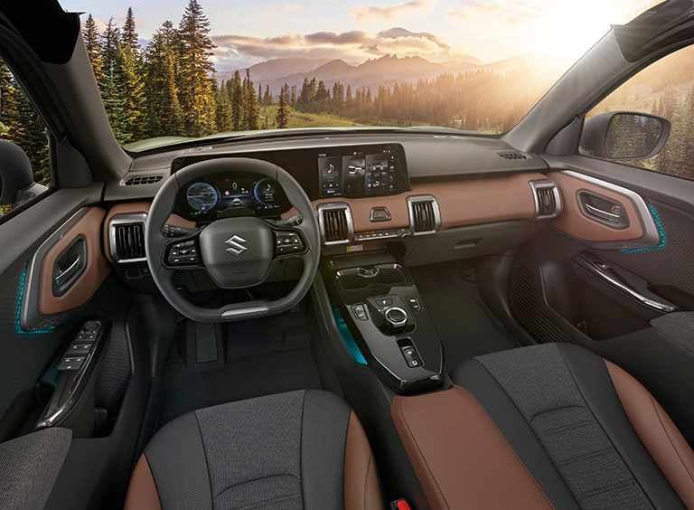 Der Innenraum des e VITARA vom Fahrersitz aus gesehen mit Fokus auf Lenkrad und Kontrollzentrum, durch die Frontscheibe sind ein sonnenbeschienener Wald und Berge zu sehen.